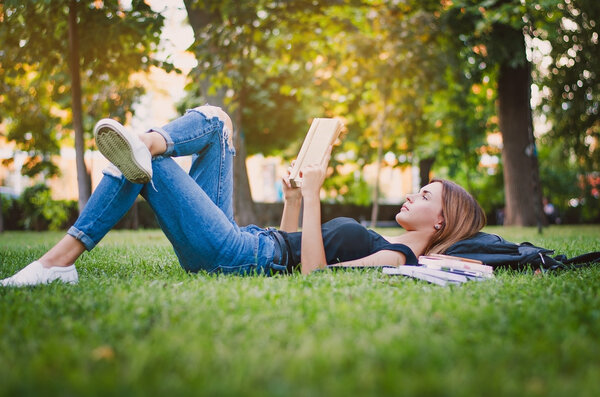 студентка в парке, лежащая на траве и читающая книгу, лежащая рядом со своим рюкзаком и стопкой тетрадей. Концепция образования
