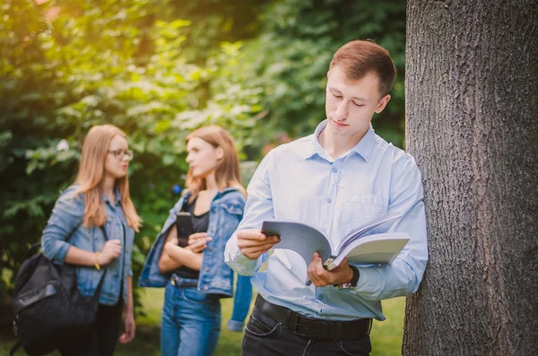 Heureux garçon souriant étudiant en bretelles, chemise et pantalon avec un livre dans le parc d'été, derrière il y a des filles et communiquer — Photo