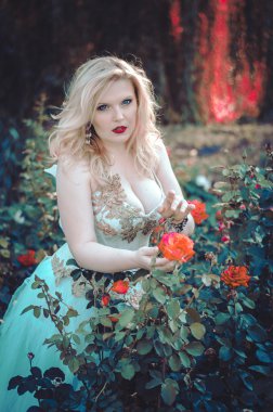 The beautiful blonde with a magnificent forms in the garden among the flowers in a long light dress. Bride on the walk