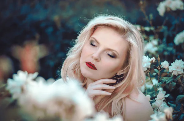 Porträt einer schönen Blondine mit geschwungener Form, roten Lippen und blauen Augen in einem hellen Kleid in einem üppigen Garten zwischen Rosen. Braut auf dem Weg — Stockfoto