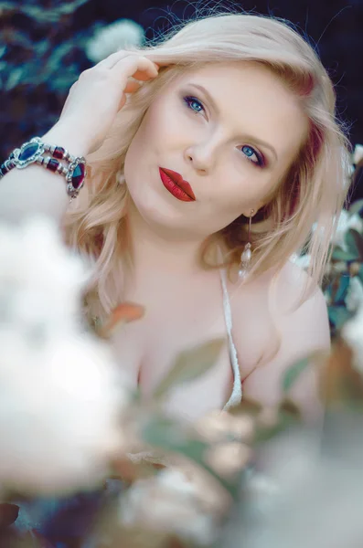 Retrato de una hermosa rubia con forma curvilínea, labios rojos y ojos azules en un vestido de luz en un exuberante jardín entre las rosas. Novia en el paseo — Foto de Stock