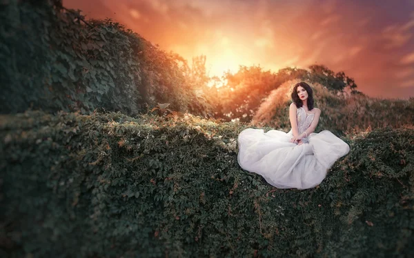 Menina bonita em um vestido branco sentado no jardim ao pôr do sol — Fotografia de Stock