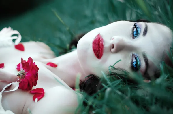 Portrait of a beautiful young brunette with red lips, blue eyes, lying on green grass with rose petals — 图库照片