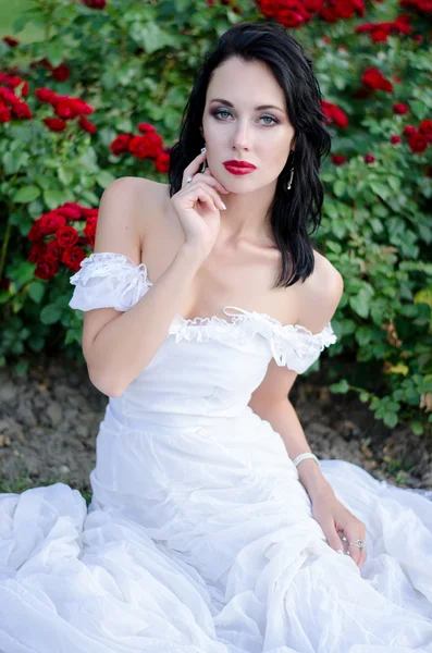 Bela jovem morena longo vestido branco sentado em um arbusto de rosas vermelhas no jardim de verão — Fotografia de Stock