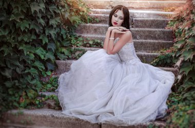 beautiful young brunette in a magnificent white dress sitting on the stairs in the garden