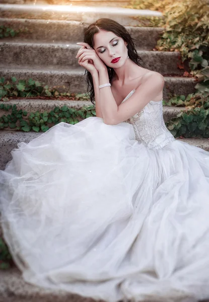 Beautiful young brunette in a magnificent white dress sitting on the stairs in the garden — Stok fotoğraf