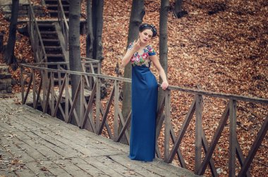 Beautiful slender brunette in a long blue dress standing on a wooden bridge in the autumn forest