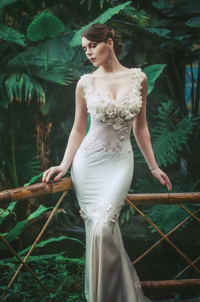 Beautiful slender young brunette in a white long slinky dress with evening hairstyle and jewelry in the jungle. Bride in the jungle. — Stok fotoğraf