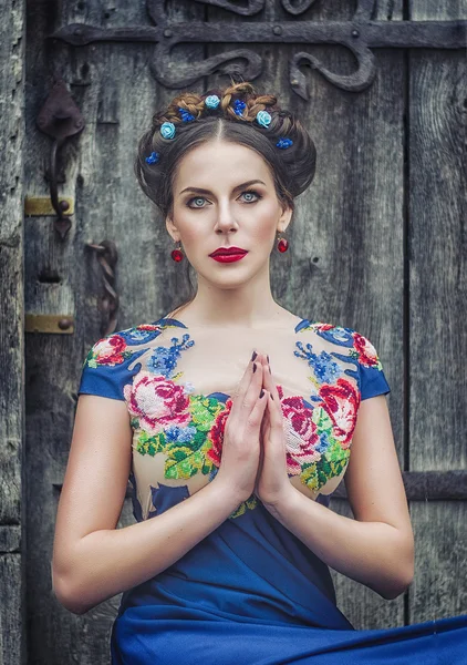 Retrato de una hermosa morena joven con un vestido azul, una corona en la cabeza, ojos azules y labios rojos sobre el fondo de una vieja puerta de madera. Deuvshka ucraniano, chica rusa — Foto de Stock