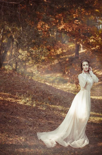 Beautiful young girl in a long gentle waving bright dress stands in the autumn forest. Bride on a walk. — Stok fotoğraf