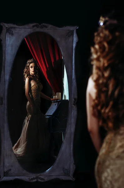 Menina bonita em um vestido amarelo longo em pé na frente de um grande espelho e olha para si mesmo no espelho . — Fotografia de Stock