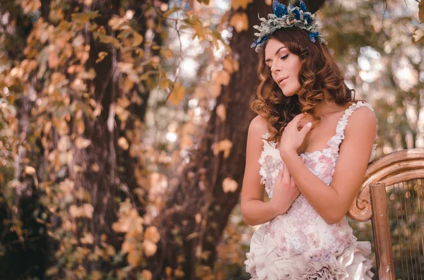 Mulher de cabelos castanhos bonita em pé em uma coroa de flores em sua cabeça e um belo vestido branco em pé em uma floresta encostada a uma harpa — Fotografia de Stock