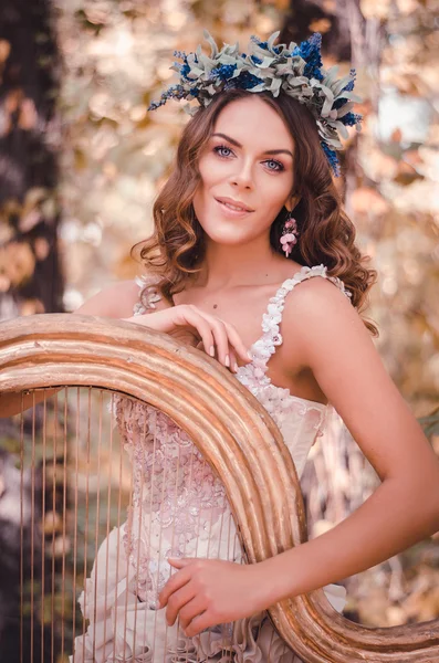 Bela mulher de cabelos castanhos com uma coroa de flores na cabeça, vestindo um vestido branco tocando harpa na floresta — Fotografia de Stock