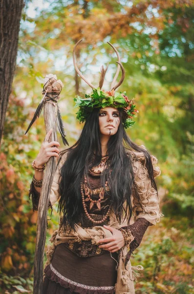 Beautiful brunette with painted face, in a flower wreath and a wooden staff in his hand, looking forward belligerently — Stock Photo, Image