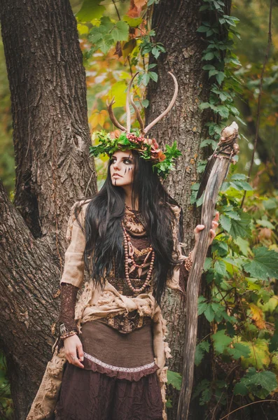 Hermosa morena en la imagen del chamán está en el bosque, sosteniendo un bastón de madera en su mano. Con una corona floral en la cabeza y cuernos — Foto de Stock