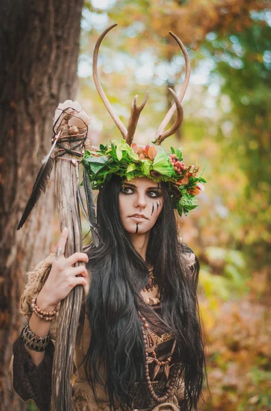 Beautiful brunette with painted face, in a flower wreath and a wooden staff in his hand, looking forward belligerently — Stock fotografie