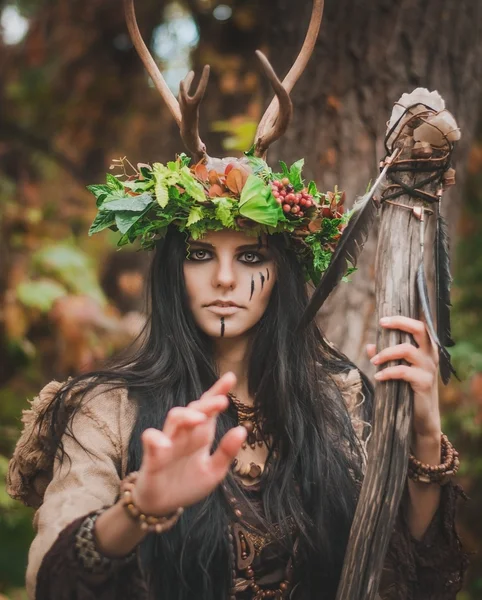 Retrato de una morena con una corona floral en la cabeza, cuernos y palo de madera en la mano, en el bosque — Foto de Stock