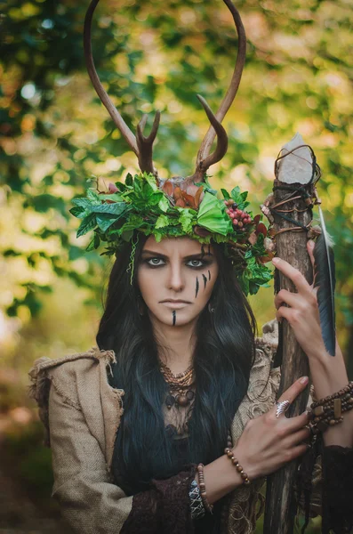 Portrait of a brunette with a floral wreath on her head, horns and wooden stick in hand, in the forest — 스톡 사진