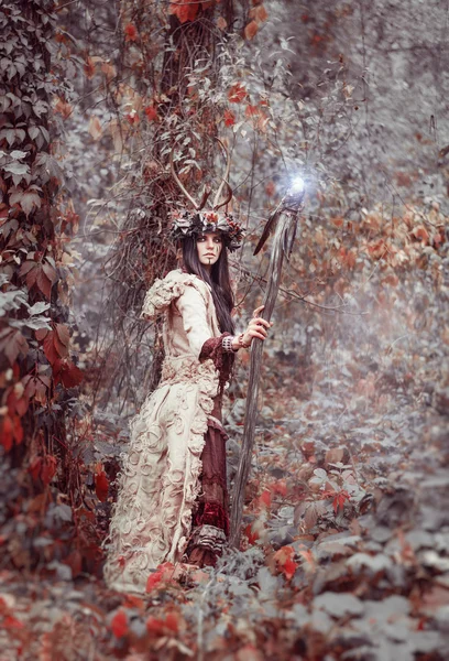 Beautiful brunette with painted face, clothes shaman, a floral wreath on her head and horns, holding a glowing wooden staff, in the woods — Zdjęcie stockowe