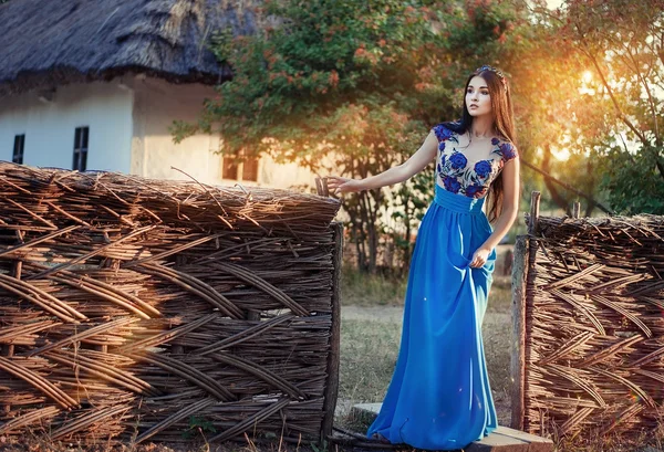 Hermosa morena delgada en un vestido largo azul con corona floral en la cabeza, de pie cerca de una vieja casa en los rayos del sol poniente —  Fotos de Stock