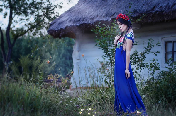 Beautiful slender brunette in a blue long dress with floral wreath on her head, standing near the old house.Ukrainian girl in village — Stock Photo, Image