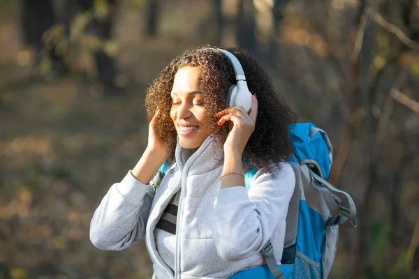 Femme afro-américaine souriante aux yeux fermés profiter de la musique tout en marchant dans la forêt avec un sac à dos. — Photo