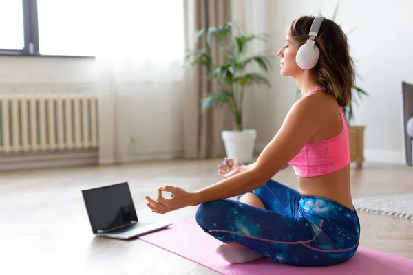 Online yoga lesson. Woman in headphones in lotus position in front of laptop monitor.