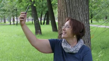 Mutlu gülümseyen son sınıf kadını şehir parkında çimlerin üzerinde otururken görüntülü arama yapıyor..