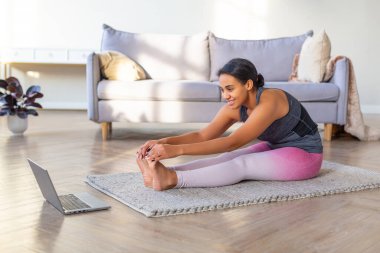 Afro-Amerikalı kadın evde esneme hareketleri yapıyor. Antrenman minderinde dizüstü bilgisayarın önünde oturuyor..