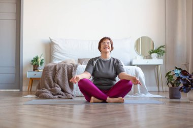 Yaşlı bir kadın evde lotus pozisyonunda meditasyon yapıyor. Sağlıklı yaşam tarzı kavramı.