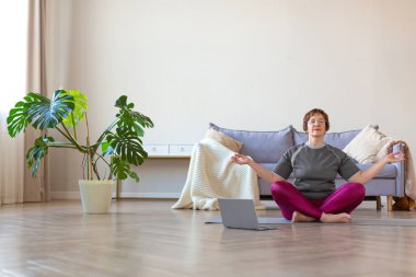 Yaşlı bir kadın evde dizüstü bilgisayar başında lotus pozisyonunda meditasyon yapıyor. Sağlıklı yaşam tarzı kavramı.