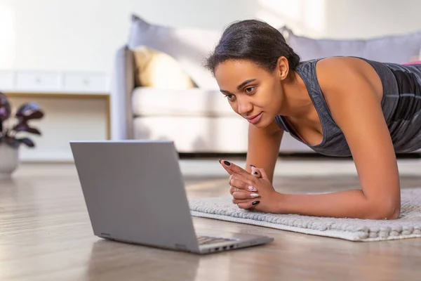 Photo of an African American woman during online workout. She stands in a plank position, following the instructions of the coach.