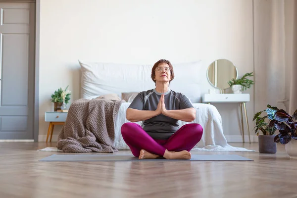 Yaşlı bir kadın evde lotus pozisyonunda meditasyon yapıyor. Sağlıklı yaşam tarzı kavramı. — Stok fotoğraf