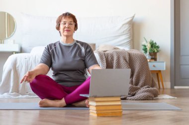 Kıdemli kadın evde dizüstü bilgisayar başında lotus pozisyonunda meditasyon yapıyor. Yaşlılıkta sağlıklı yaşam biçimi kavramı.