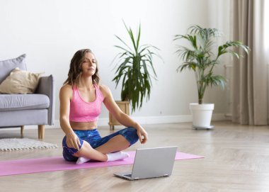 Genç bir kadın internette özel ders kullanarak meditasyon yapıyor. Laptop monitörünün önünde gözleri kapalı oturuyor..