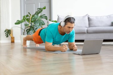 Orta yaşlı, kulaklıklı bir adamın tahta egzersizi yaparken fotoğrafı. İnternetteki dersleri takip ediyor..