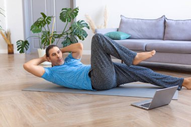 Orta yaşlı bir adam karın kaslarını çalıştırıyor, dizüstü bilgisayarda spor videosu izlerken karın kaslarını çalıştırıyor..