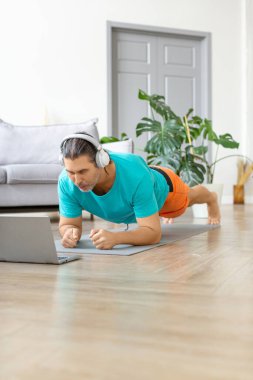 Orta yaşlı, kulaklıklı bir adamın tahta egzersizi yaparken fotoğrafı. İnternetteki dersleri takip ediyor. Dikey fotoğraf.