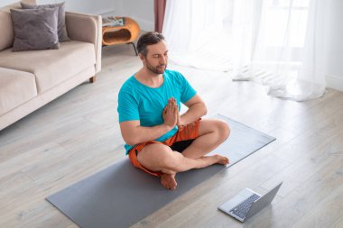 Orta yaşlı bir adam dizüstü bilgisayarın önünde meditasyon yapıyor..