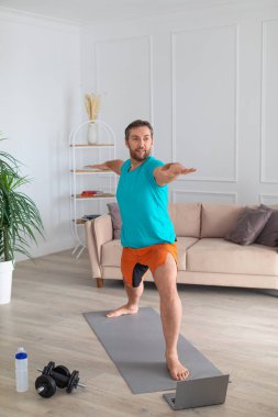 Orta yaşlı bir adam evde online öğretmenlik yapıyor. Dizüstü bilgisayarın önünde denge egzersizi yapıyor. Dikey fotoğraf.