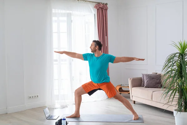 Man working out at home with an online tutorial. He performs a balance exercise in front of a laptop monitor.