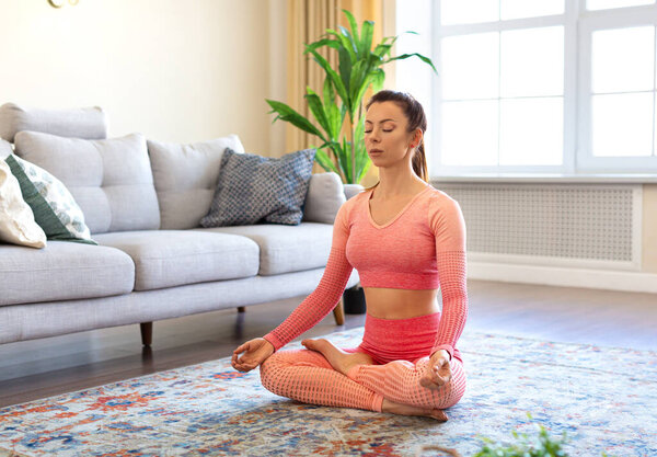Side view of a woman is meditating at home. She sits in lotus position on the floor with her eyes closed and practices yoga meditation.