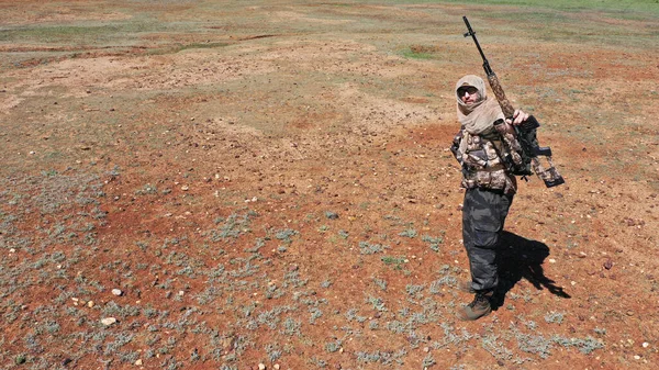 Foto de um atirador no deserto. Ele se levanta, olha para a câmera e mostra sua espingarda. — Fotografia de Stock