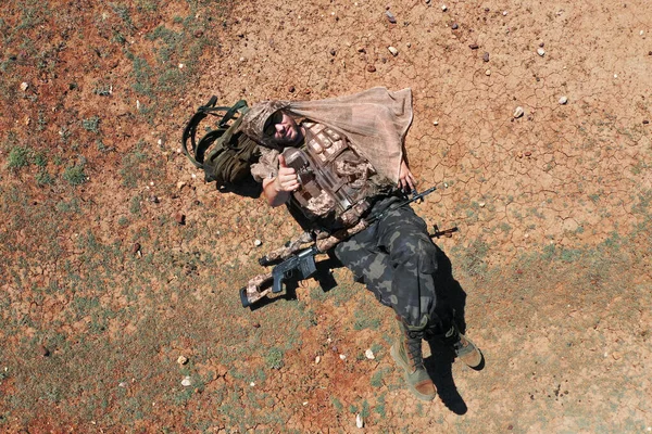 Visão superior - um atirador soldado enquanto descansa. Ele está deitado no deserto vermelho-quente e descansa. Ele mostra os polegares para a câmara.. — Fotografia de Stock