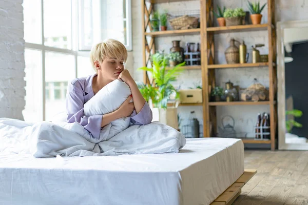 Sad blonde woman sitting on the bed in the morning and hugging her pillow. She didnt get enough sleep.