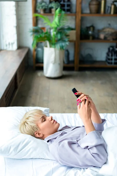 수직 사진. 여자가 휴대폰을 들고 침대에 누워 있습니다. 일어나서 소셜 미디어 게시물을 보고 있었어요. — 스톡 사진