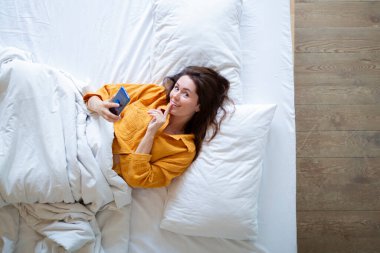 Top view of smiling young woman lying in bed with smartphone.