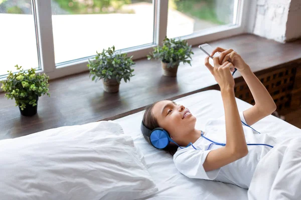 10 살난 여자 애가 침대에서 음악을 듣습니다. 헤드폰을 통해 음악을 듣고 스마트폰으로 온라인 앱을 이용해 음악을 듣는다. — 스톡 사진