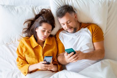 Orta yaşlı çiftin akıllı telefonlarıyla yatakta yatışının üst görüntüsünü kapat..