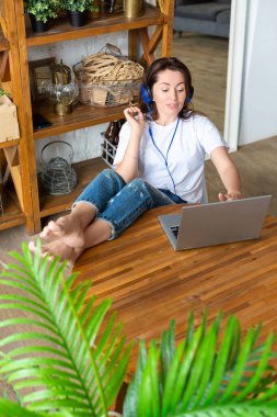 Vertical photo of a happy smiling woman in blue headphones in front of a laptop monitor at home. She puts her feet on the table and vividly discusses the latest gossip with her friend. clipart